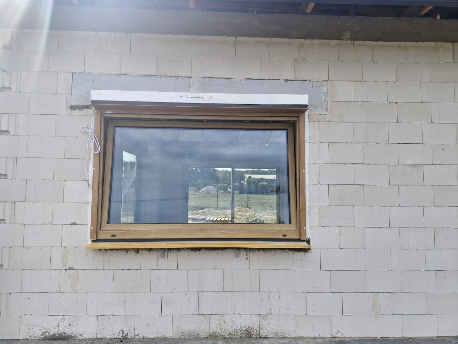 Montaż okna z roletą zewnętrzną w surowym stanie, widoczny mur z bloczków, częściowo otynkowane nadproże, niezabezpieczona roleta zwinięta nad oknem, widok na zewnątrz przez szybę.