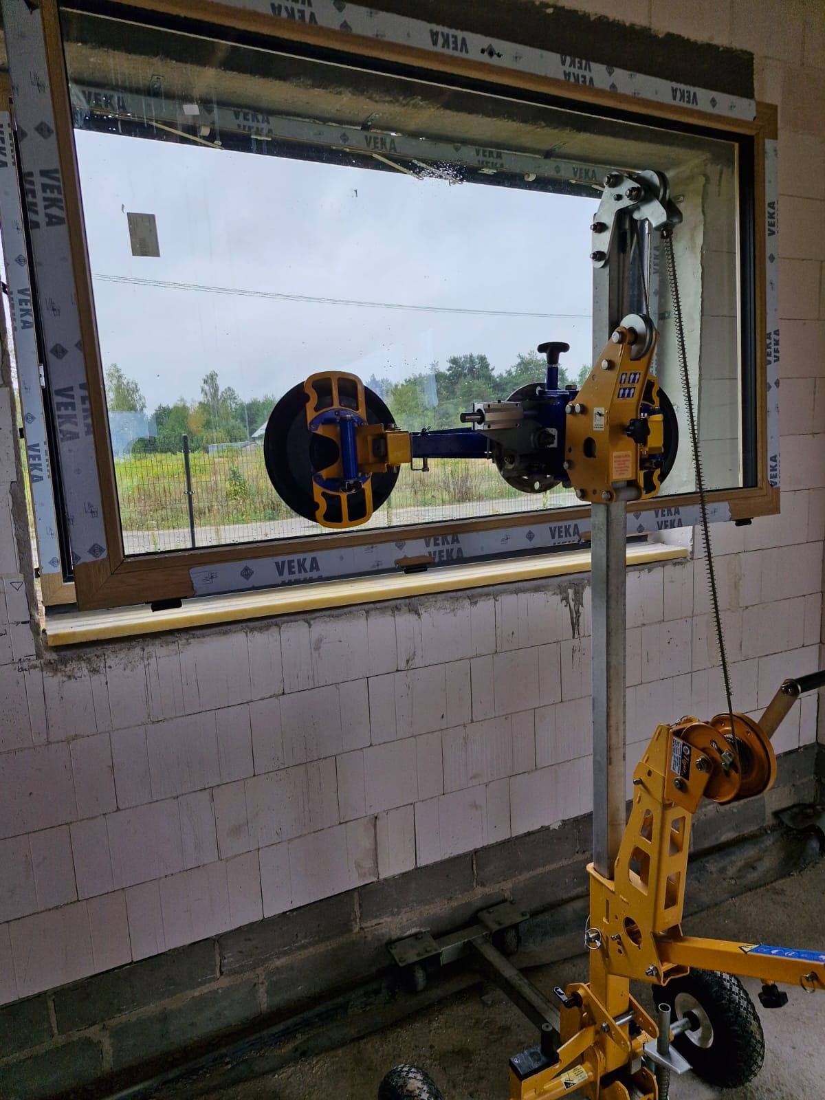 Montaż okna z użyciem specjalistycznego podnośnika z przyssawkami, widoczne profile okienne marki VEKA i surowy mur z betonu komórkowego.