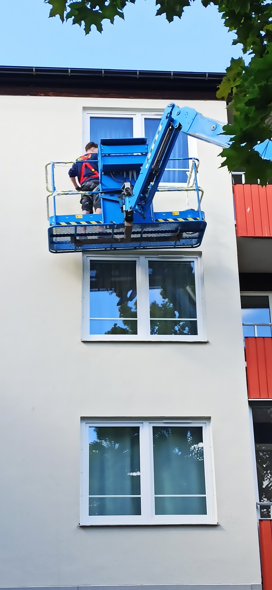 Pracownik w niebieskim podnośniku koszowym montuje lub naprawia okno PCV w białej ramie na elewacji budynku.