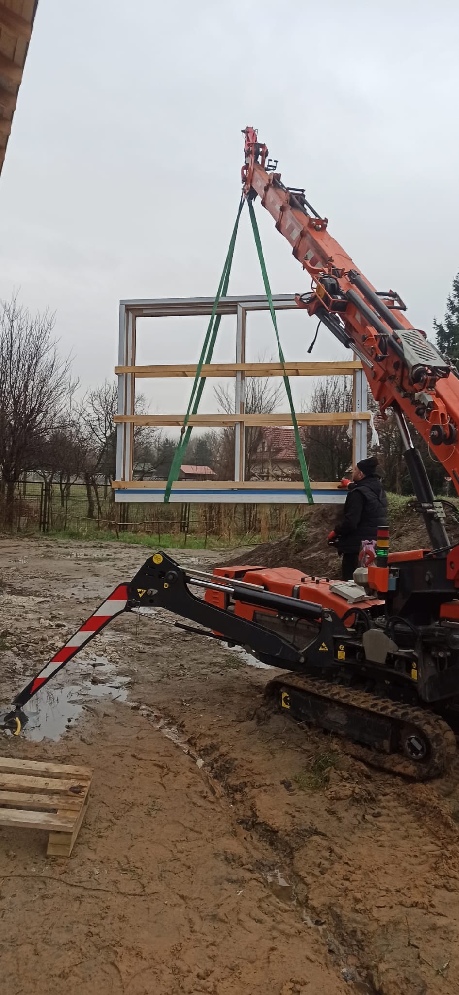 Montaż dużego okna przez dźwig na błotnistym terenie, widoczny pracownik w czarnej kurtce i operator dźwigu.