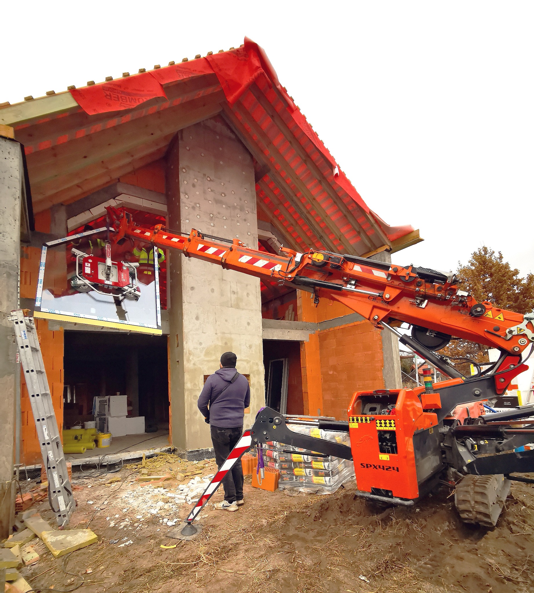 Montaż okna z wykorzystaniem specjalistycznego dźwigu przyssawkowego SPX424 na budowie domu jednorodzinnego w stanie surowym otwartym, widoczny pracownik nadzorujący proces.
