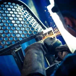 Spawanie metalowej konstrukcji krzesła z perforowanym siedziskiem; widoczne iskry i spawacz w masce ochronnej, pracujący w warsztacie.