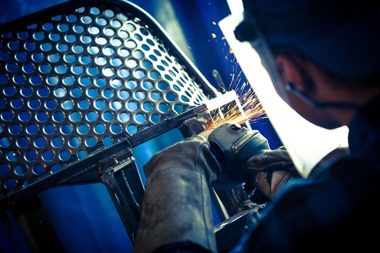 Spawanie metalowej konstrukcji krzesła z perforowanym siedziskiem; widoczne iskry i spawacz w masce ochronnej, pracujący w warsztacie.