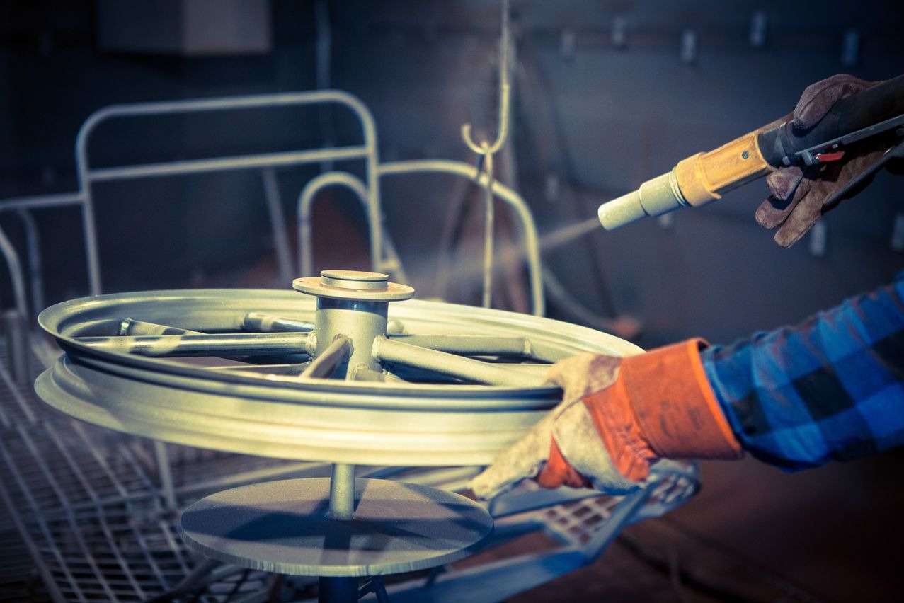 Metalowe koło motocyklowe na obrotowym stojaku jest malowane proszkowo przez pracownika w rękawicach ochronnych i niebiesko-czarnej koszuli.