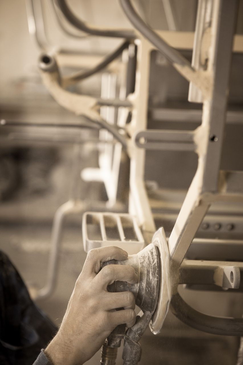 Szlifowanie metalowej konstrukcji za pomocą szlifierki oscylacyjnej, widoczne opiłki, surowy stan wykończenia, szaro-beżowa kolorystyka.