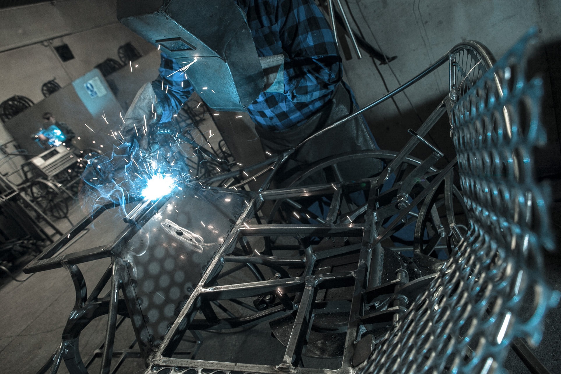 Spawanie metalowej konstrukcji z siatką, widoczne iskry i spawacz w masce ochronnej i niebieskiej koszuli w kratę, w tle inne metalowe elementy.