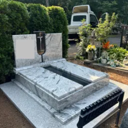 Jasnoszary, granitowy pomnik z krzyżem wkomponowanym w płytę główną, umieszczony na świeżo usypanej ziemi na cmentarzu, z białym samochodem dostawczym w tle.