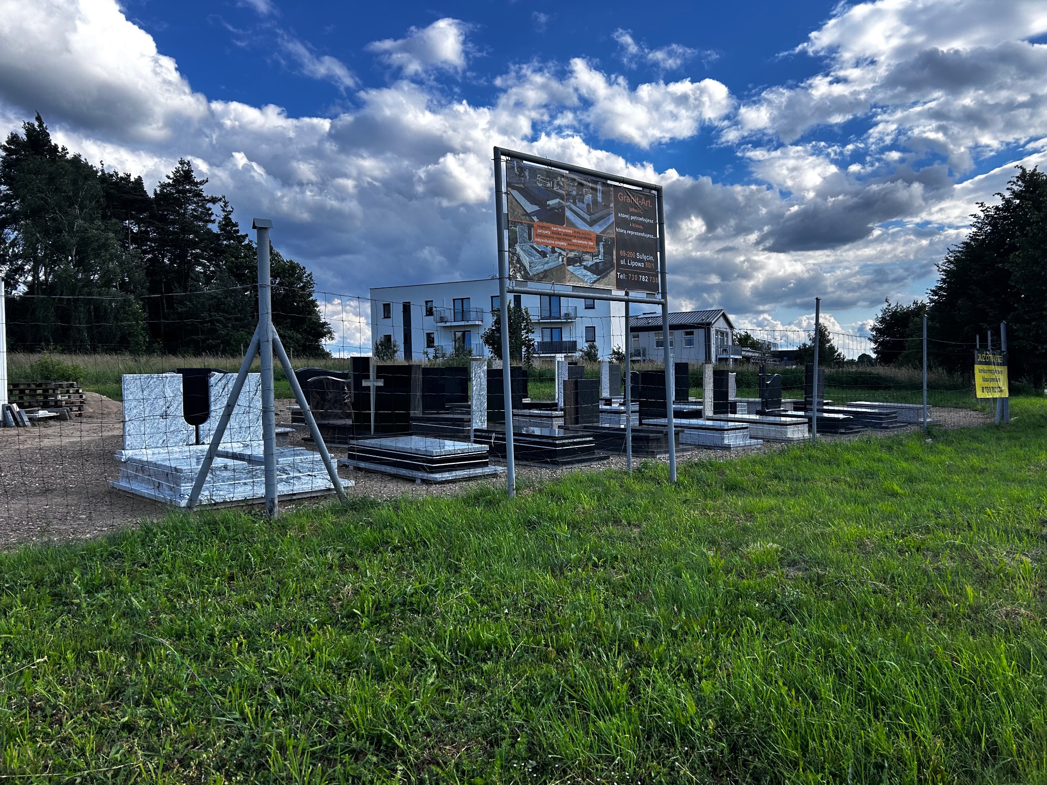 Ekspozycja nagrobków z ciemnego kamienia na trawiastym terenie, częściowo owiniętych folią ochronną, z widoczną tablicą reklamową firmy kamieniarskiej na tle nowoczesnego budynku i błękitnego nieba...