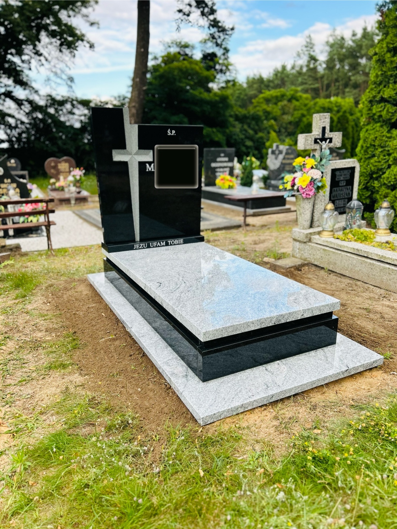 Nowoczesny, czarno-szary pomnik z krzyżem na cmentarzu, z napisem 'Jezu ufam Tobie', otoczony zielenią i innymi nagrobkami.