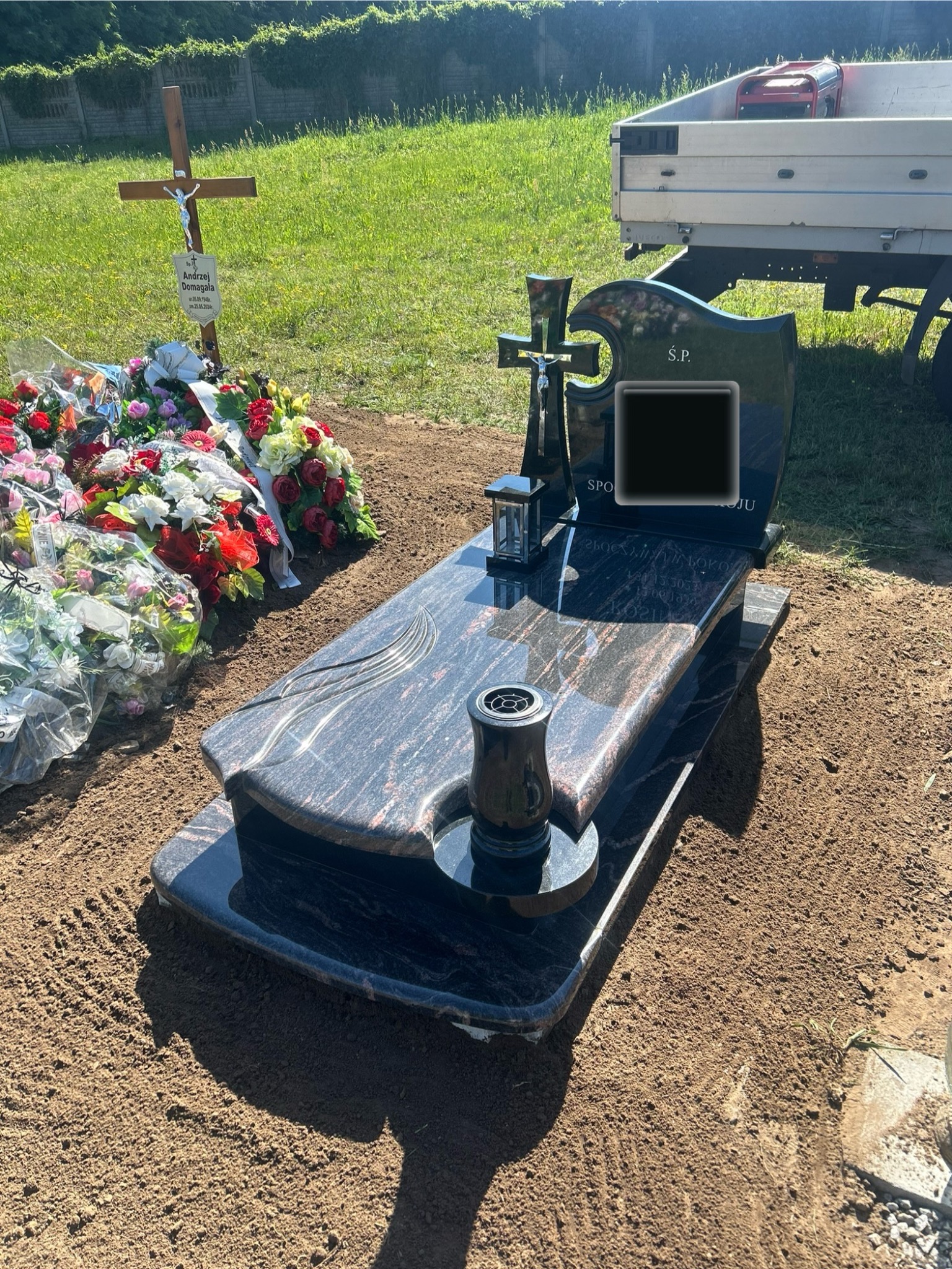 Czarny, polerowany nagrobek z krzyżem i miejscem na znicz, otoczony świeżymi kwiatami, na świeżo usypanej ziemi, z drewnianym krzyżem w tle i fragmentem przyczepy samochodowej.