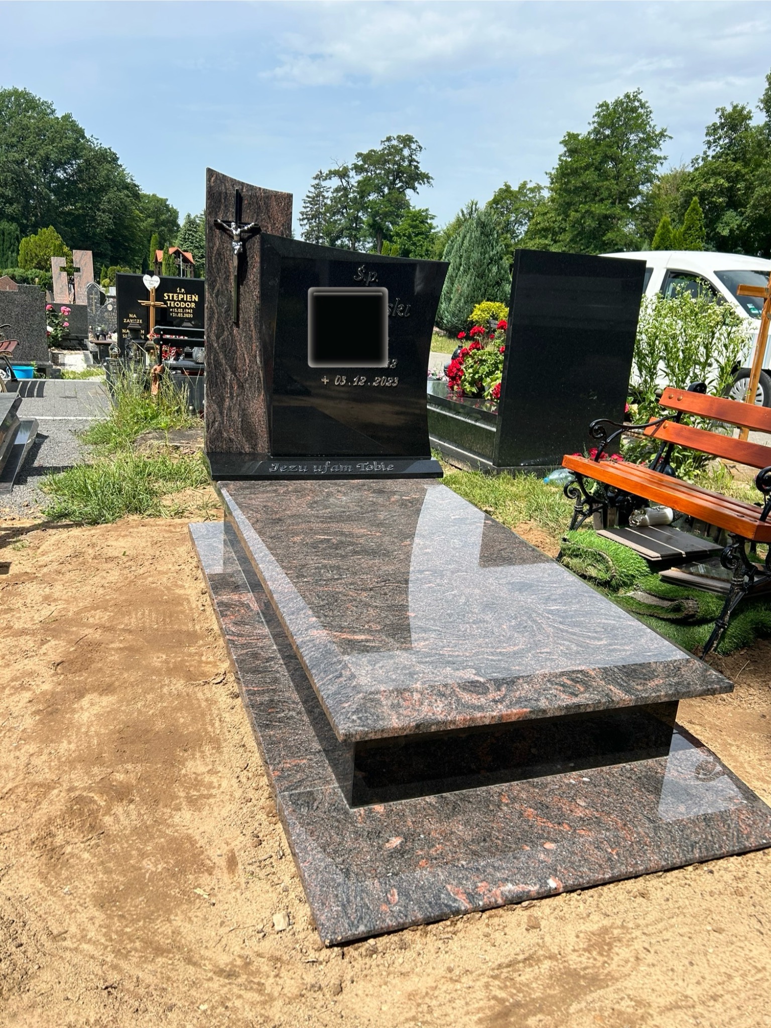 Wypolerowany, dwupoziomowy pomnik z ciemnego, ziarnistego granitu z krzyżem, na tle cmentarza z ławką i innymi nagrobkami.