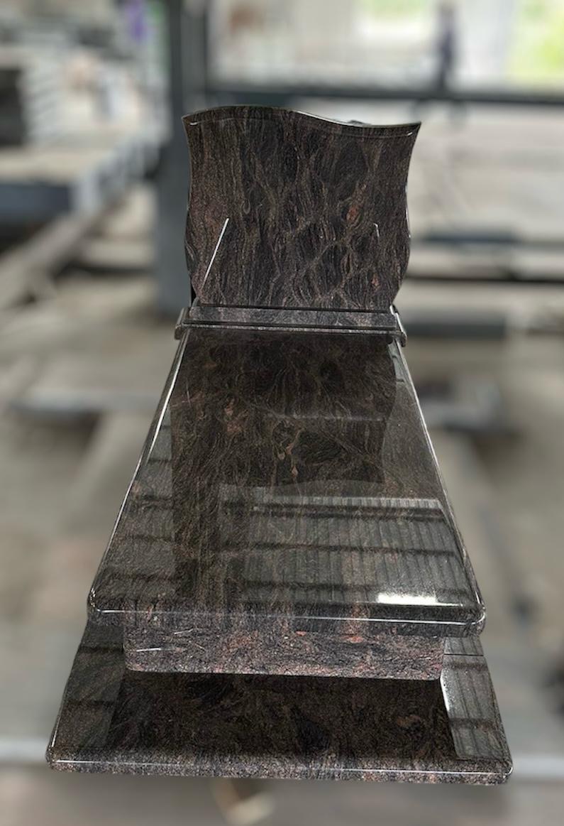 Polerowany, gotowy nagrobek z ciemnego, wzorzystego kamienia w zakładzie kamieniarskim, prezentujący unikalne ułożenie warstw i faliste wykończenie górnej części.