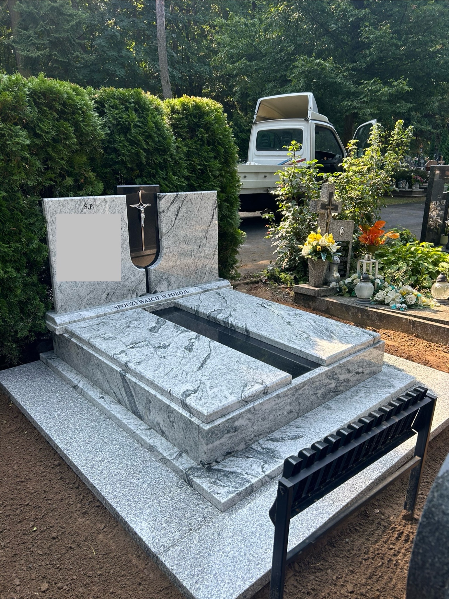 Jasnoszary, granitowy pomnik z krzyżem wkomponowanym w płytę główną, umieszczony na świeżo usypanej ziemi na cmentarzu, z białym samochodem dostawczym w tle.