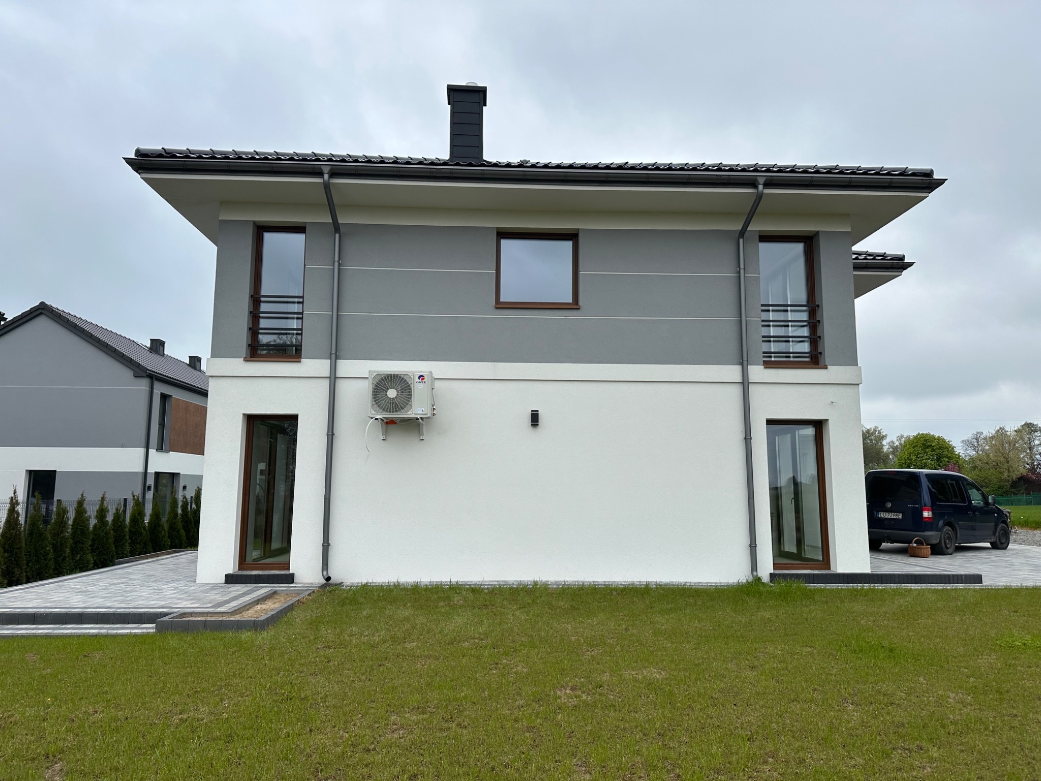 Dwukondygnacyjny dom w Lublinie z zamontowaną jednostką klimatyzacji na białej ścianie parteru, widoczny z zewnątrz na tle zielonego trawnika i szarego nieba.