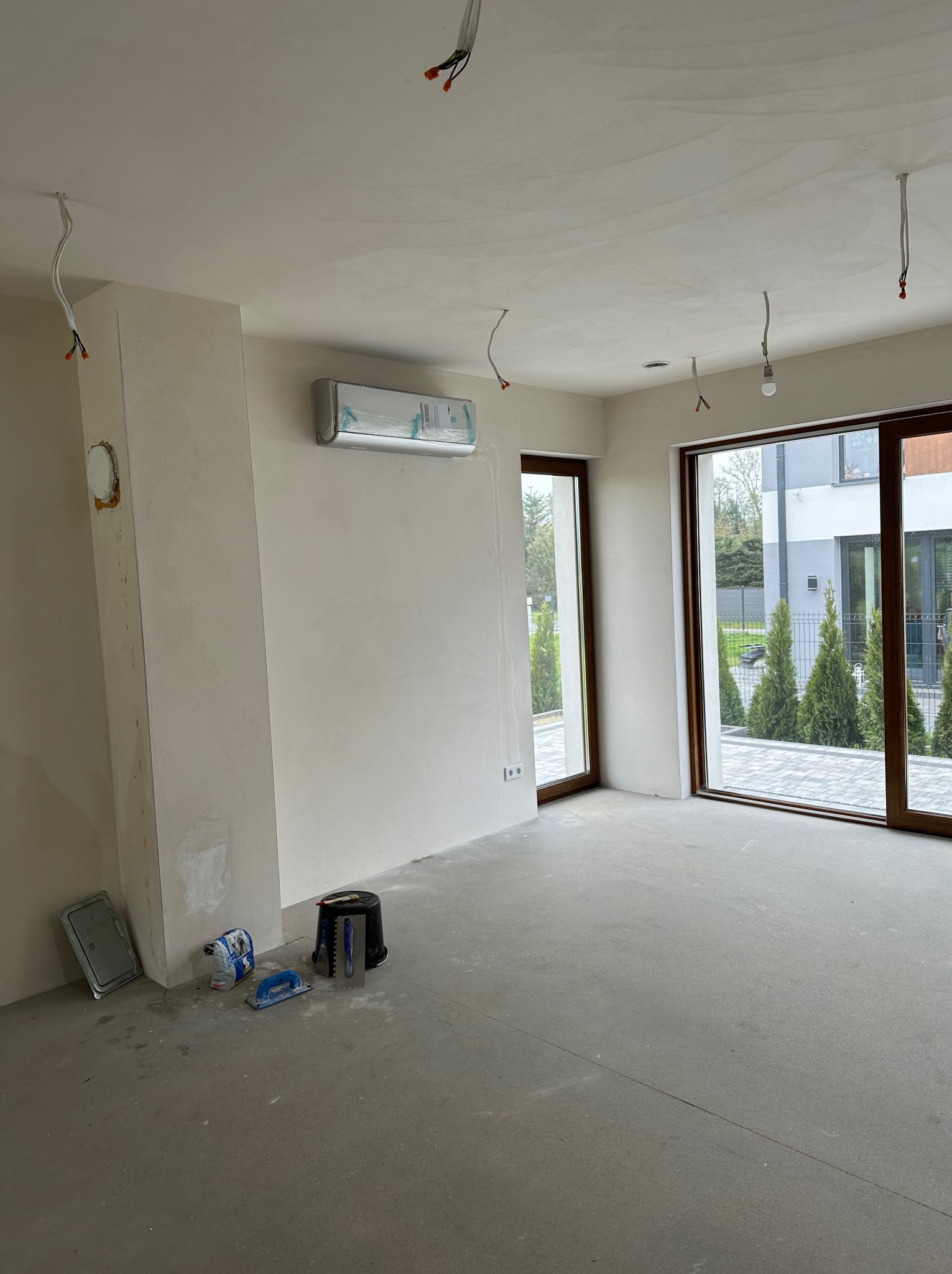 Wnętrze w trakcie remontu z zamontowanym klimatyzatorem, widoczne narzędzia malarskie i wykończeniowe, surowe ściany i podłoga, przewody elektryczne wystające z sufitu.