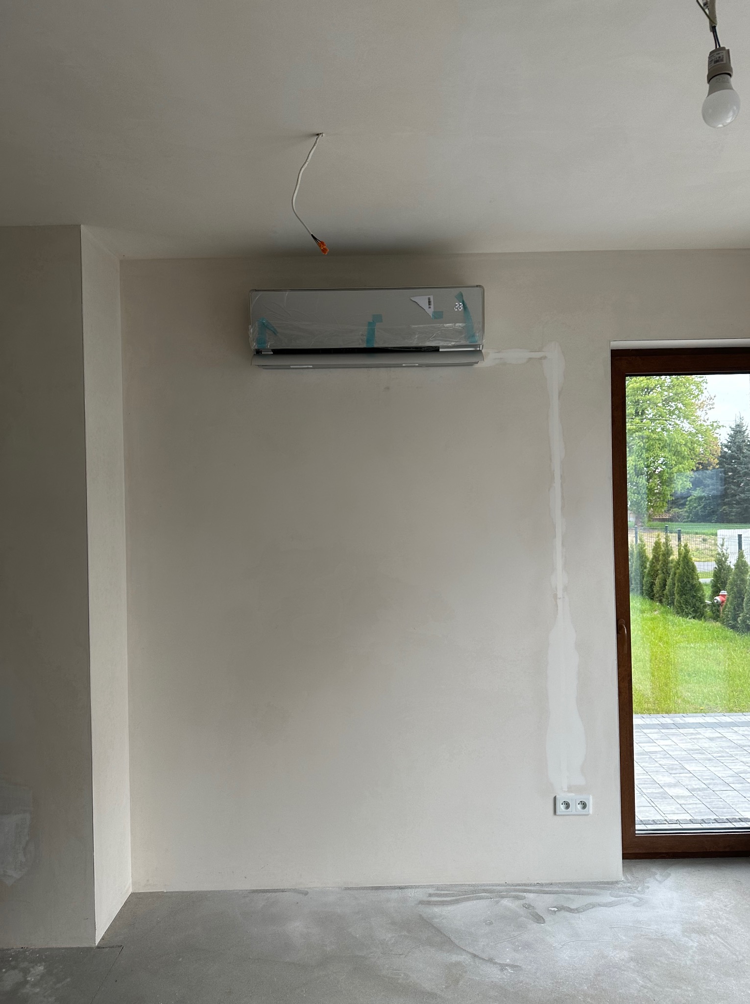 Nowo zamontowany klimatyzator ścienny, jeszcze w folii ochronnej, w surowym wnętrzu z widocznymi śladami szpachlowania na ścianie i betonową podłogą. Przez okno widoczny zadbany ogród.