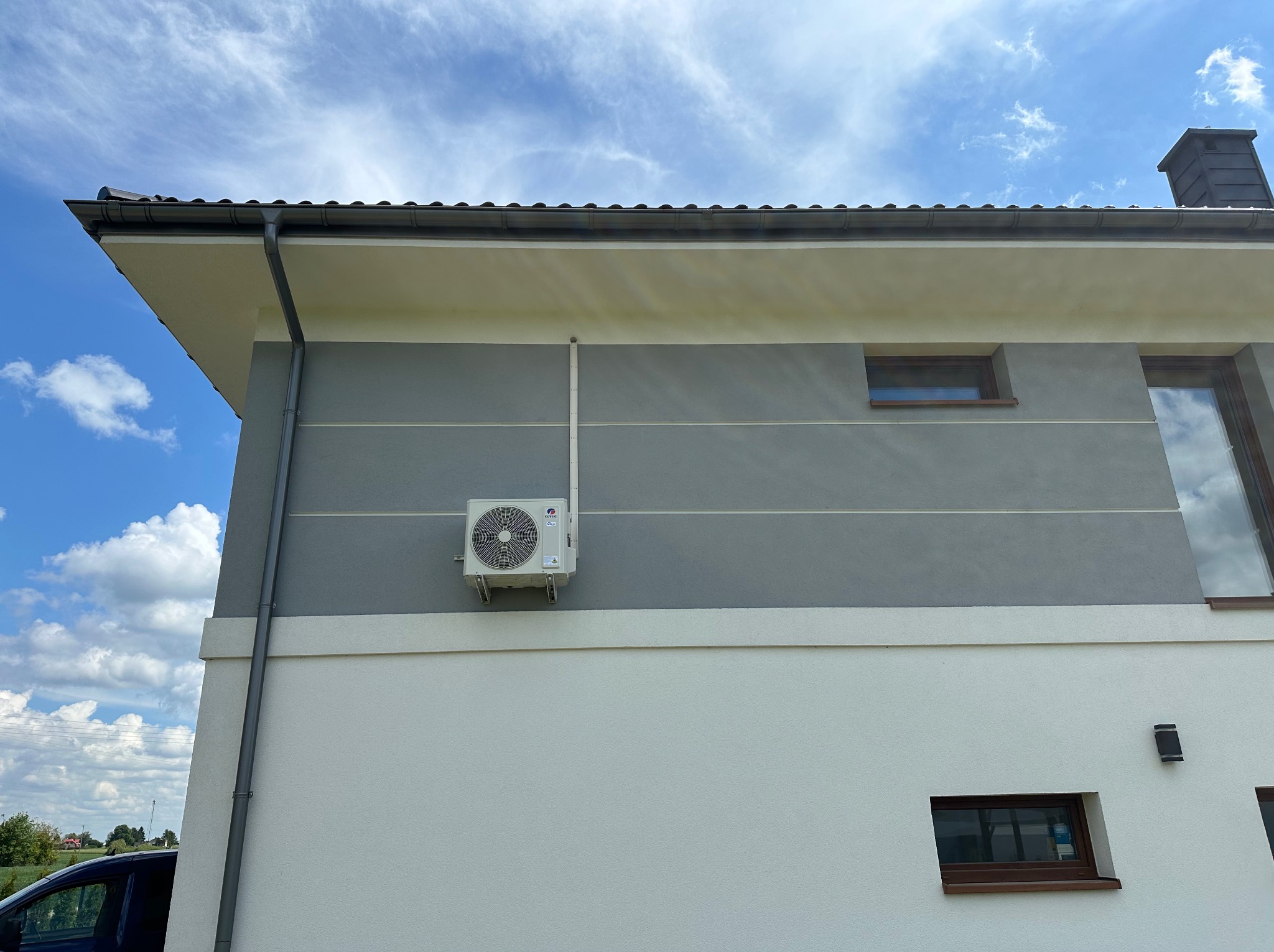 Zewnętrzna jednostka klimatyzacji marki Gree zamontowana na szarej ścianie nowoczesnego domu z widocznymi oknami i rurą odprowadzającą skropliny, na tle błękitnego nieba z chmurami.