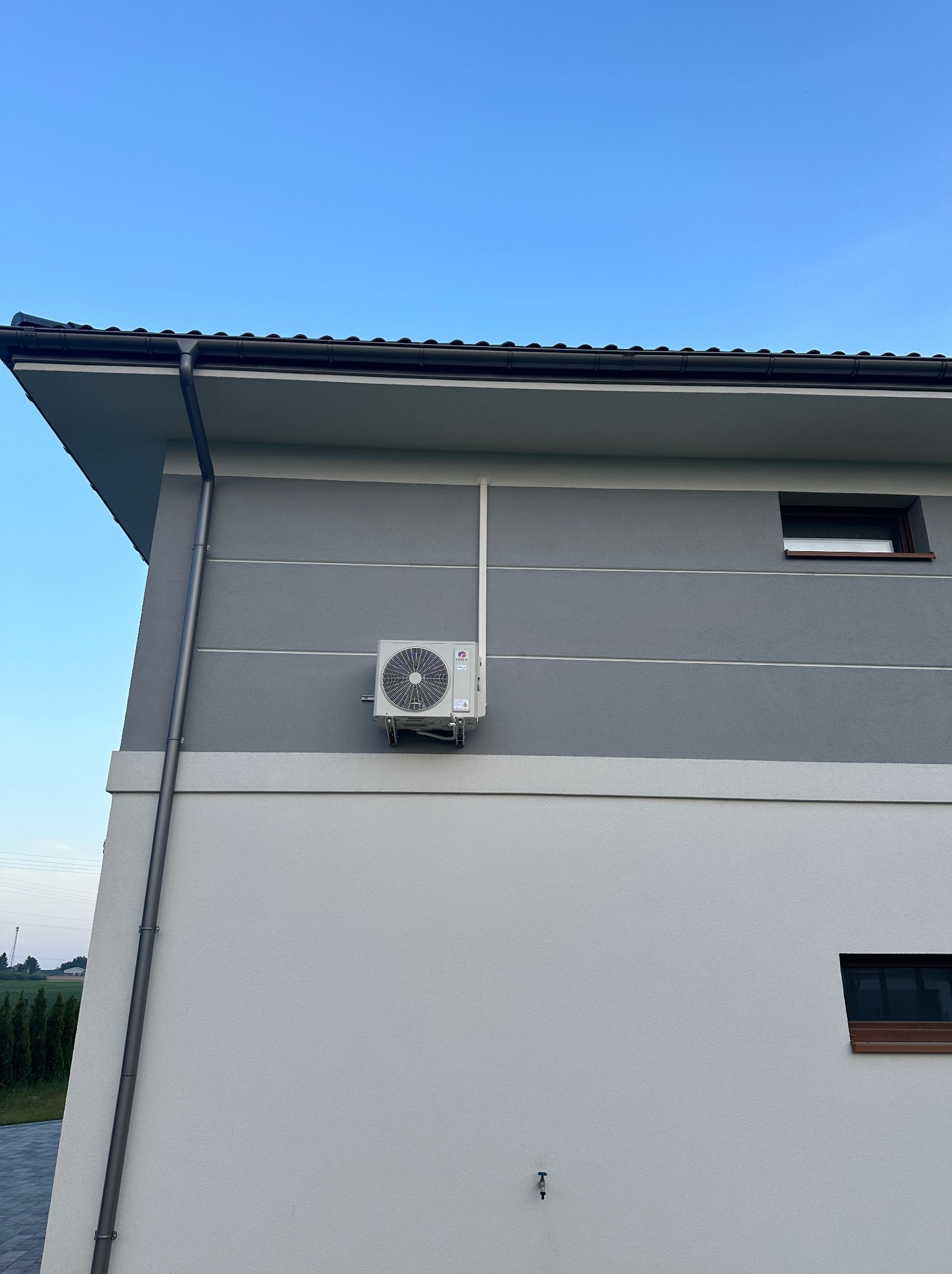 Zewnętrzna jednostka klimatyzacji zamontowana na dwukolorowej elewacji nowoczesnego domu z widocznymi rurami odprowadzającymi skropliny i instalacją elektryczną.