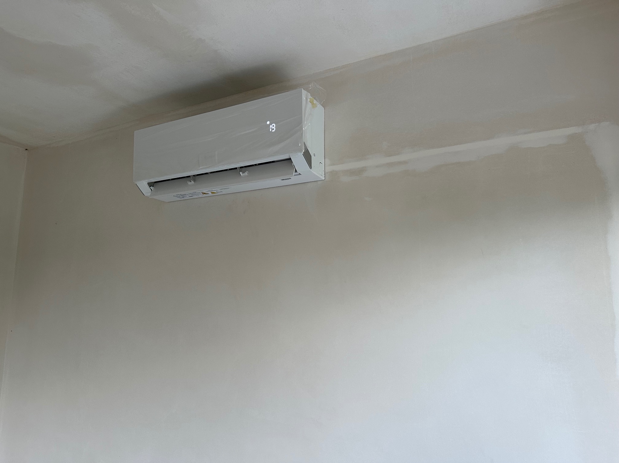 Nowo zamontowany, biały klimatyzator na ścianie w trakcie remontu, z widoczną folią ochronną i wyświetlaczem temperatury ustawionym na 19 stopni Celsjusza.