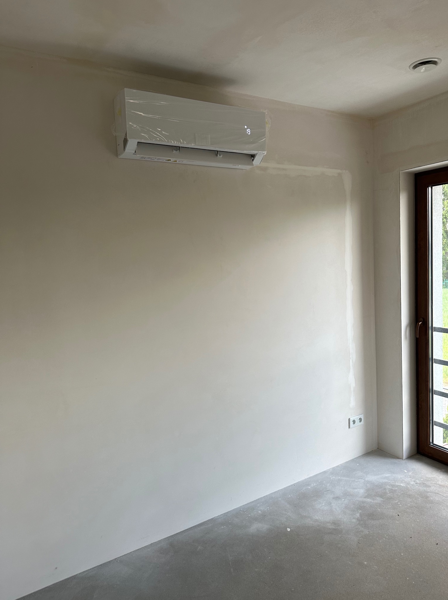 Klimatyzator ścienny, jeszcze w folii ochronnej, zamontowany w surowym pomieszczeniu z widocznymi śladami po szpachlowaniu i gładzi, z oknem balkonowym w tle.