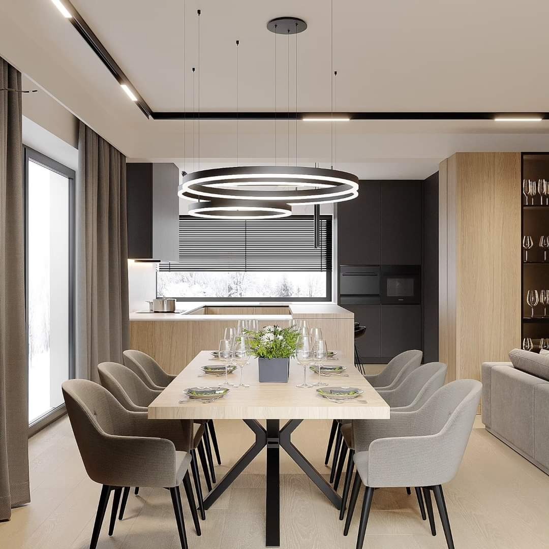 Nowoczesna jadalnia z drewnianym stołem na metalowej podstawie, szarymi krzesłami, okrągłą lampą wiszącą i widokiem na kuchnię z oknem.