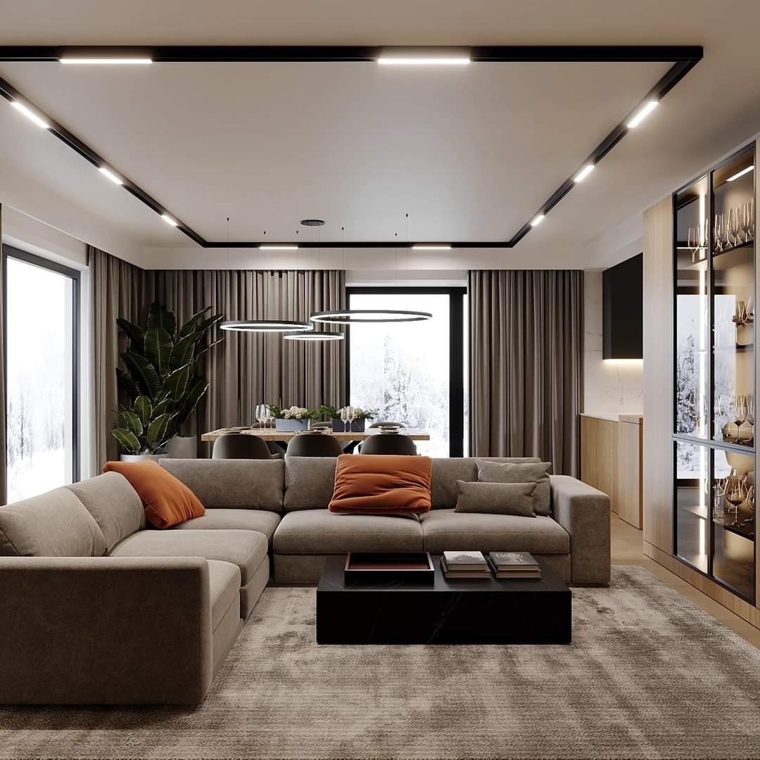 Nowoczesny salon z narożną sofą w odcieniach beżu, pomarańczowe poduszki, czarny stolik kawowy, dywan z długim włosiem, okrągłe lampy wiszące i przeszklona witryna z dekoracyjnym szkłem.