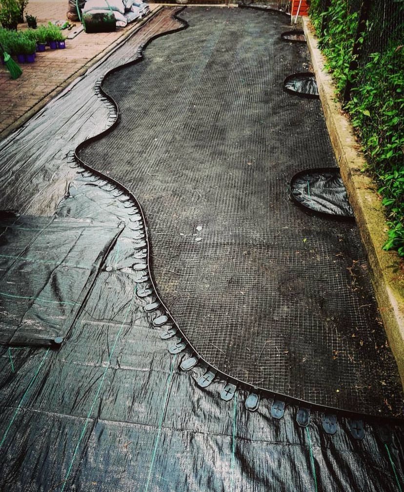 Przygotowanie terenu pod ogród: rozłożona czarna agrowłóknina z wyciętymi okręgami, obrzeże trawnikowe z plastikowymi kołkami, widoczne worki z ziemią i sadzonki w tle.