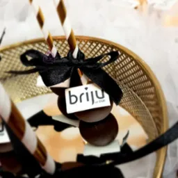 Czekoladowe lizaki z logo firmy 'brijú od 1920' na złotym talerzu, ozdobione czarną kokardą i papierowymi słomkami w złote paski.