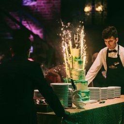 Każdego Słodkiego Agnieszka Pasiecka-Kwiatkowska - Trzypiętrowy tort w odcieniach zieleni, z fontannami iskier, na stole z zielonym obrusem, kelner w tle, stosy talerzy obok.