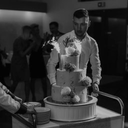 Każdego Słodkiego Agnieszka Pasiecka-Kwiatkowska - Czarno-białe zdjęcie przedstawia trzy poziomowy tort weselny z różami i fontannami iskier, transportowany na wózku przez mężczyznę w białej koszuli, w tle widoczni goście.