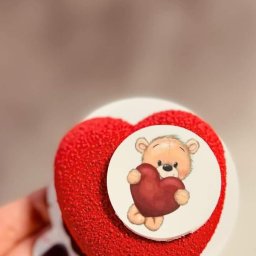 Każdego Słodkiego Agnieszka Pasiecka-Kwiatkowska - Czerwone ciastko w kształcie serca z pluszowym misiem trzymającym serce na białym okrągłym tle, trzymane w dłoni z ciemnym lakierem do paznokci.