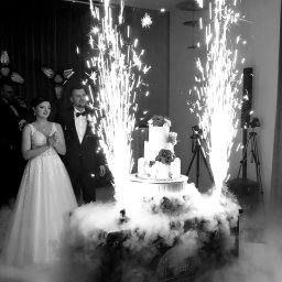 Każdego Słodkiego Agnieszka Pasiecka-Kwiatkowska - Czarno-białe zdjęcie nowożeńców stojących obok trzypiętrowego tortu weselnego udekorowanego różami, otoczonego dymem i fontannami iskier.