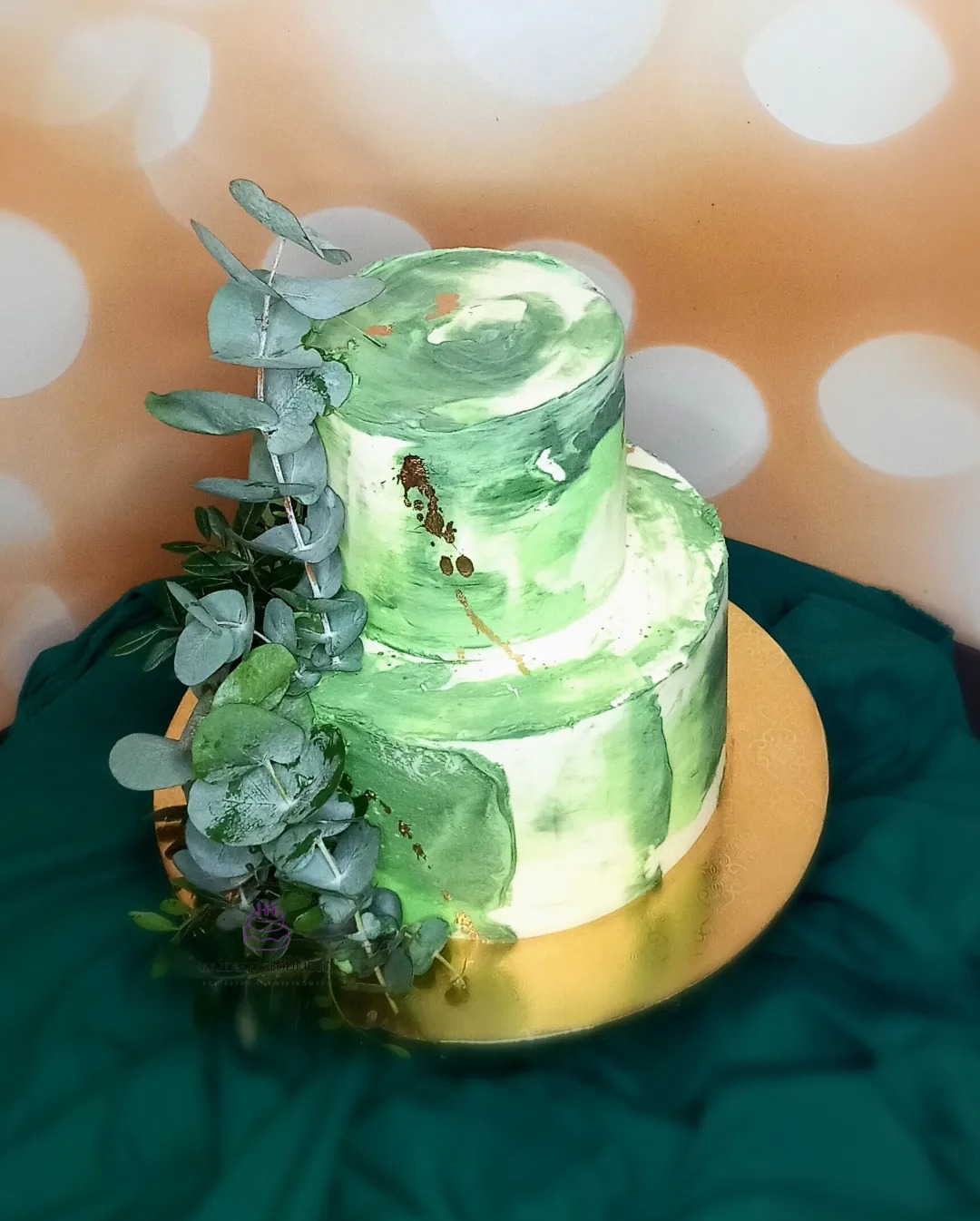 Dwupoziomowy tort w odcieniach zieleni z marmurkowym wzorem, udekorowany gałązkami eukaliptusa, na złotej paterze i zielonej tkaninie.