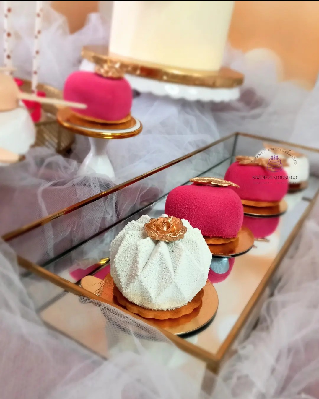 Eleganckie mini desery: białe, teksturowane kulki z złotymi kwiatami i różowe serca z złotymi akcentami, ułożone na lustrzanej tacy z złotymi brzegami, z tortem w tle i dekoracyjną tkaniną.