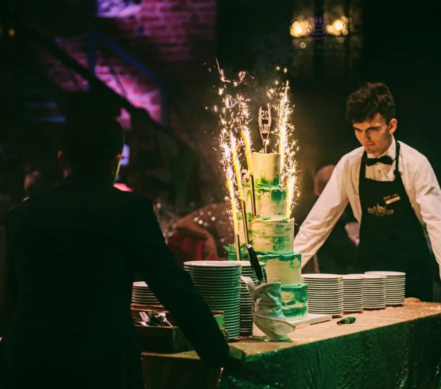 Trzypiętrowy tort w odcieniach zieleni, z fontannami iskier, na stole z zielonym obrusem, kelner w tle, stosy talerzy obok.