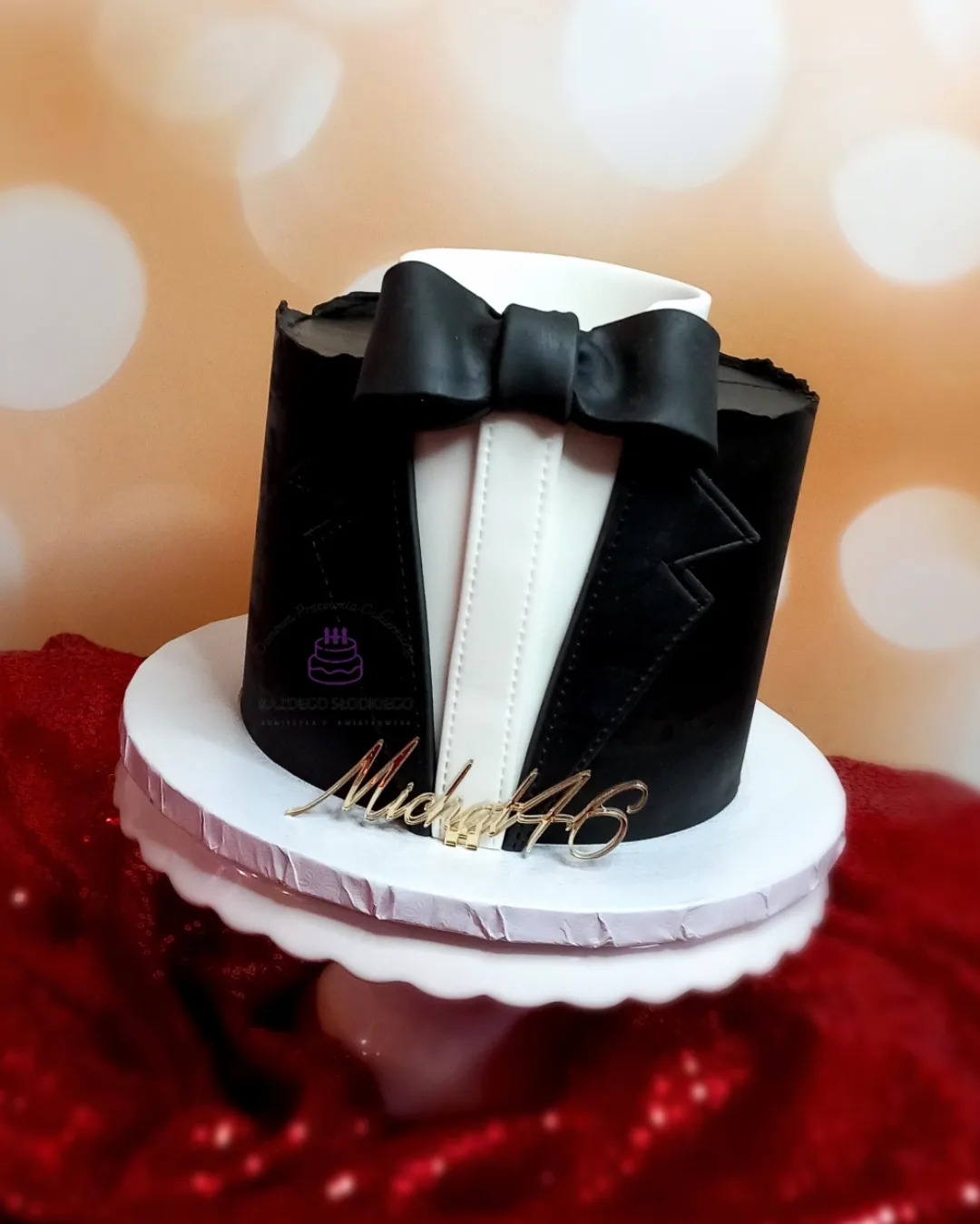 Tort urodzinowy w kształcie fraka z czarną muchą i napisem 'Michał 46' na białym stojaku, na tle bokeh.