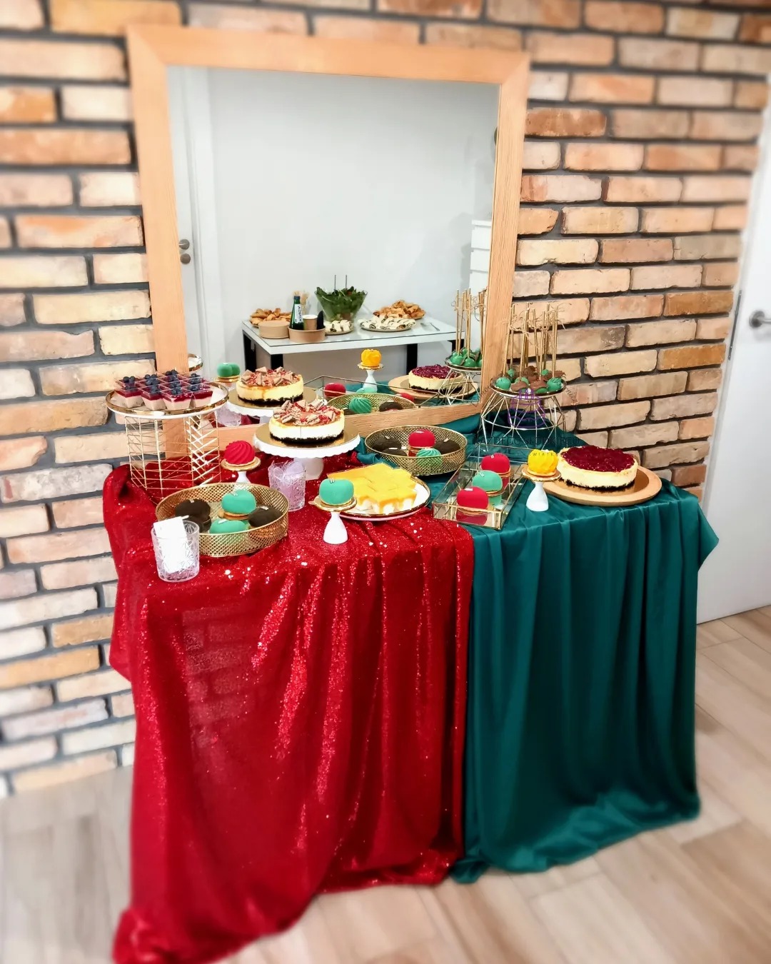 Stół z deserami: mini torciki, ciasta i ciasteczka w kolorowych dekoracjach na złotych paterach, ustawione na czerwonym i zielonym błyszczącym obrusie, z lustrem w tle.