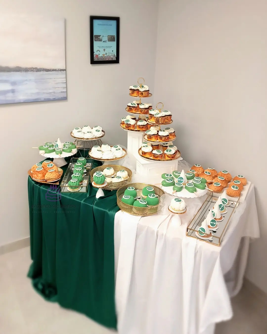 Stół z zielono-białym obrusem, na którym znajdują się różne słodkości, takie jak babeczki, ciasteczka i bezy, niektóre z zielonymi dekoracjami, ustawione na złotych paterach i tacach, w tle obraz...