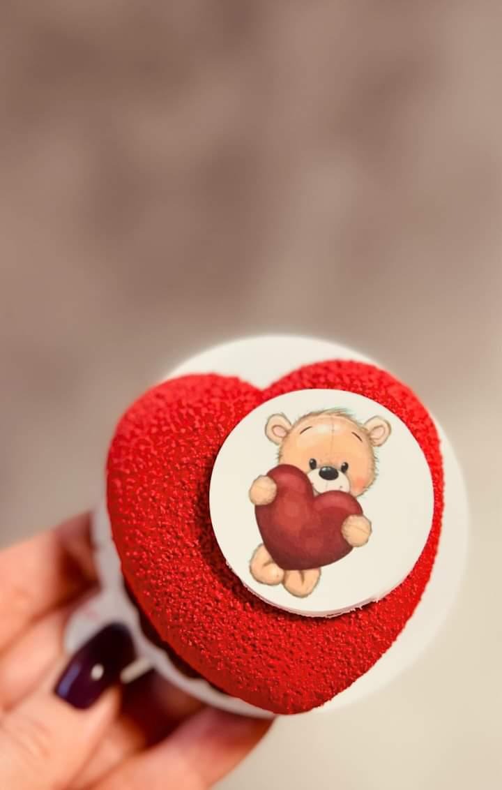 Czerwone ciastko w kształcie serca z pluszowym misiem trzymającym serce na białym okrągłym tle, trzymane w dłoni z ciemnym lakierem do paznokci.