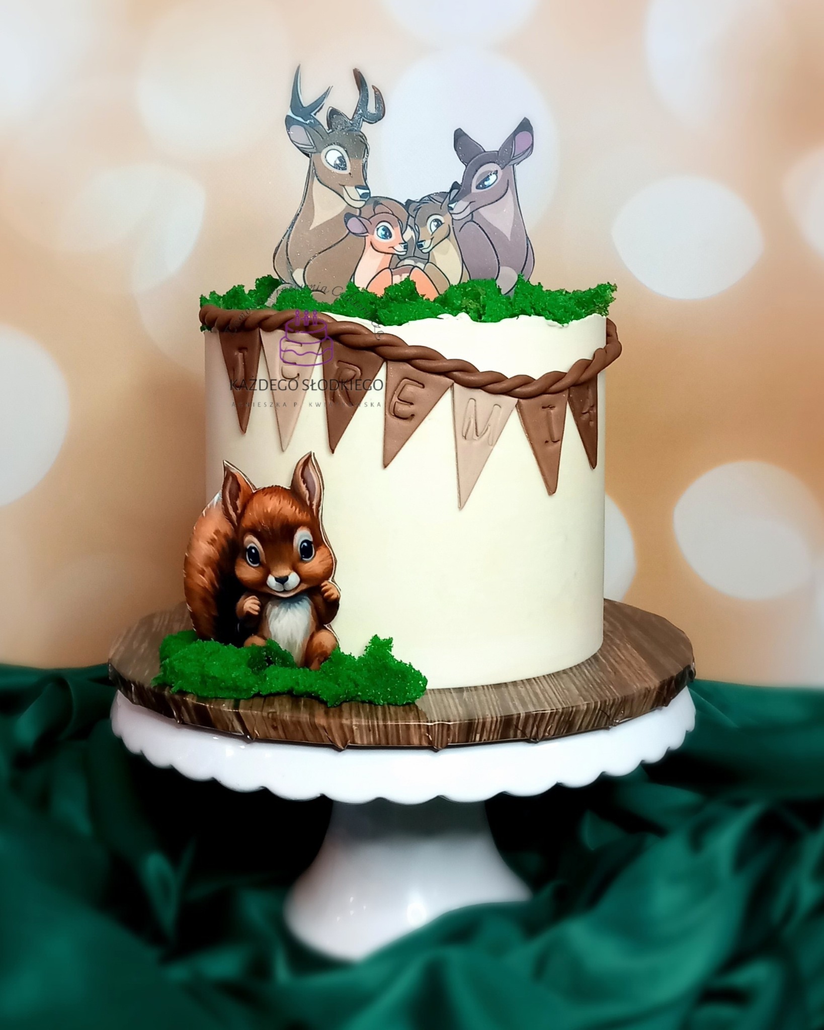 Tort urodzinowy z motywem leśnych zwierząt, Bambi i wiewiórka, na białym stojaku, dekorowany brązowym lukrem imitującym gałęzie i zielonym lukrem imitującym trawę, z napisem na chorągiewkach.