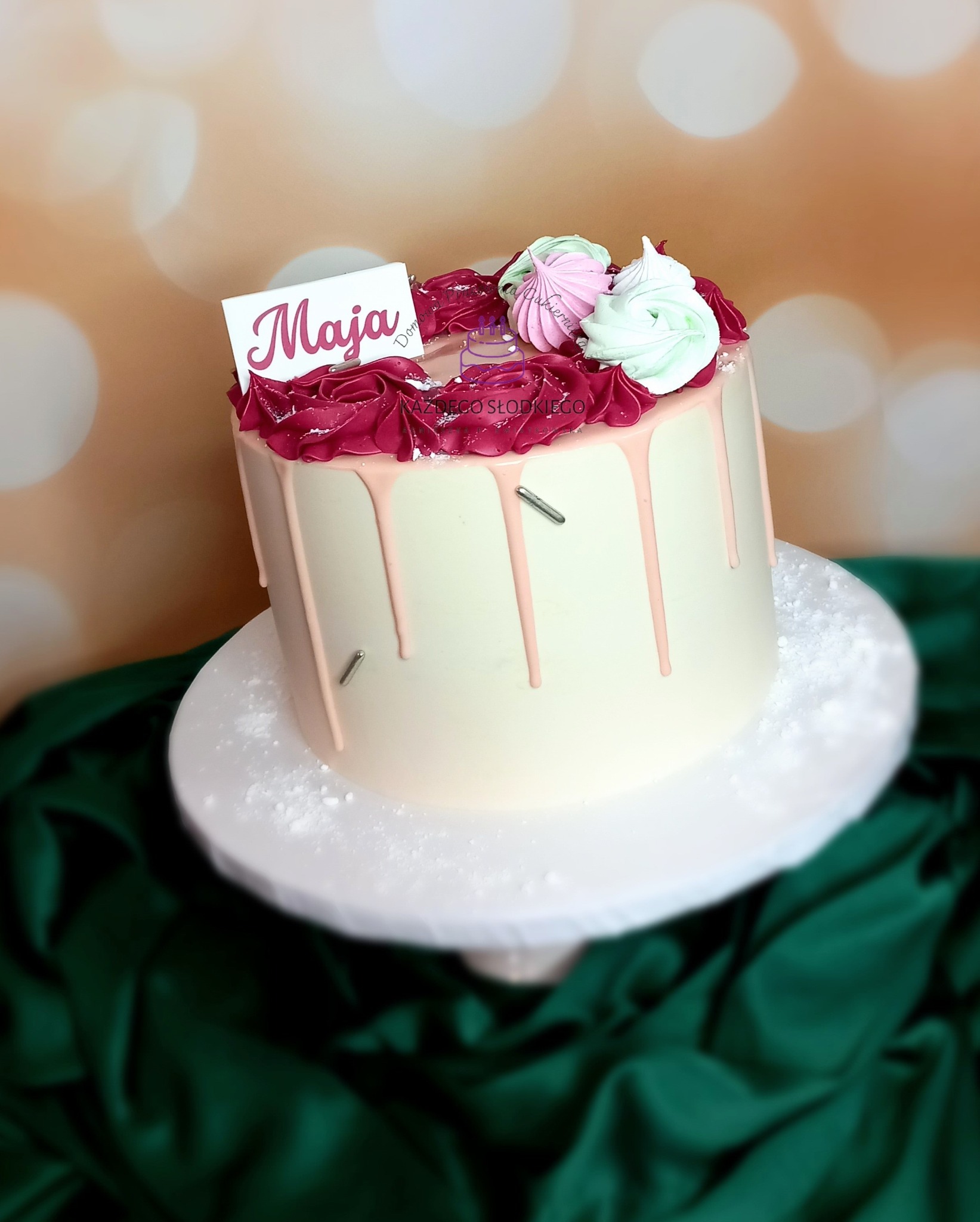 Tort urodzinowy z białym kremem, polewą w odcieniach różu i dekoracjami z bezy, ozdobiony tabliczką z napisem 'Maja', umieszczony na białym stojaku na tle zielonej tkaniny i bokeh.