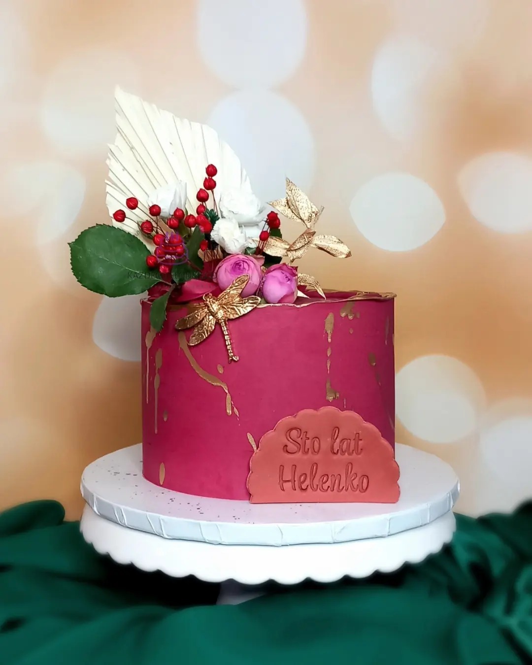 Tort urodzinowy w kolorze fuksji, ozdobiony złotymi akcentami, kwiatami, liśćmi i napisem 'Sto lat Helenko', umieszczony na białym stojaku na tle bokeh.