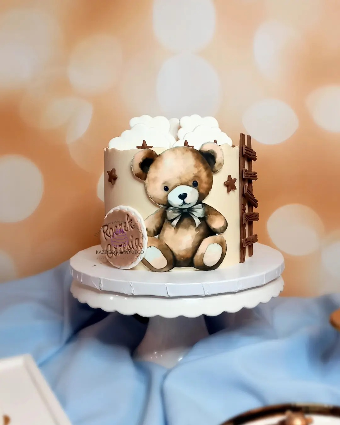 Tort urodzinowy z motywem misia, dekorowany chmurkami, gwiazdkami i czekoladową drabinką, umieszczony na białym stojaku.