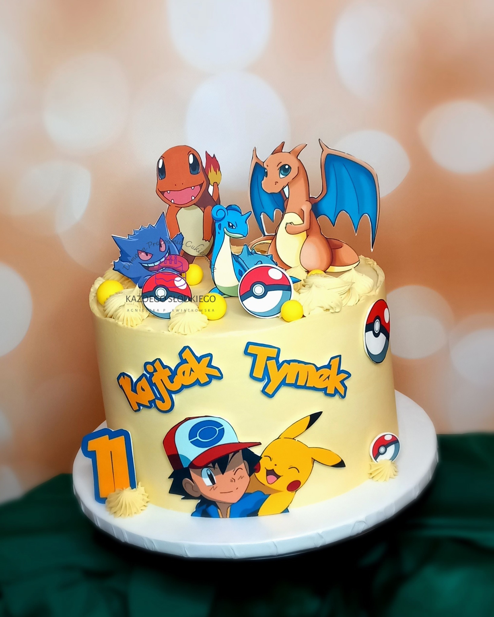 Tort urodzinowy w stylu Pokemon z figurkami Charmandera, Charizarda, Laprasa i Gengara, udekorowany imionami Kajtek i Tymek oraz numerem 11, na tle bokeh.