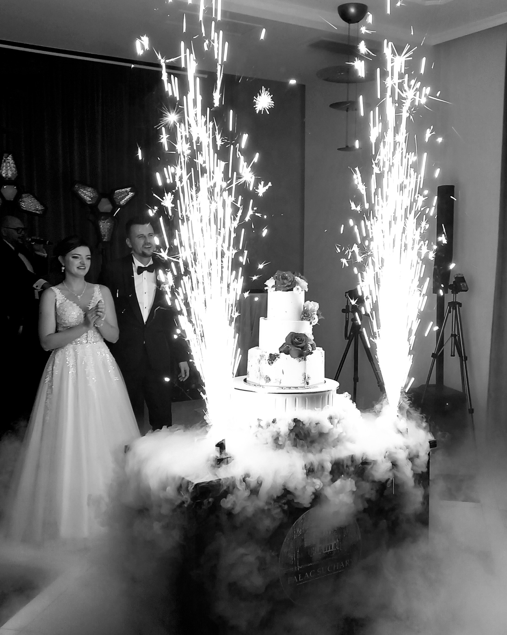 Czarno-białe zdjęcie nowożeńców stojących obok trzypiętrowego tortu weselnego udekorowanego różami, otoczonego dymem i fontannami iskier.