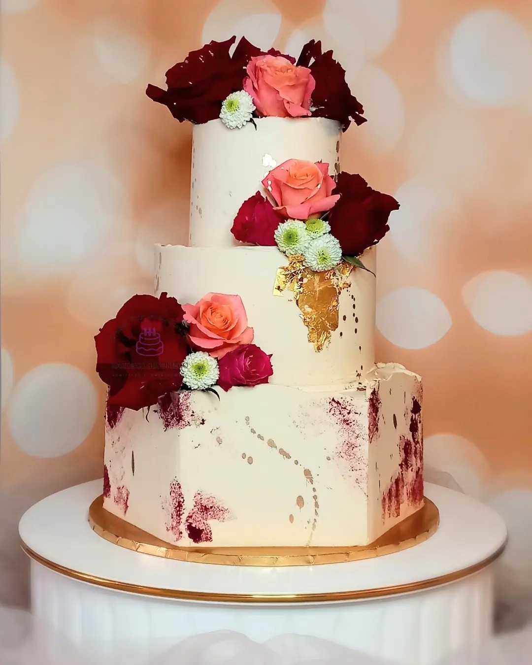 Elegancki, trzypiętrowy tort weselny z kremowym lukrem, zdobiony świeżymi czerwonymi i różowymi różami oraz złotymi płatkami, umieszczony na białym podeście z obramowaniem w kolorze złota.