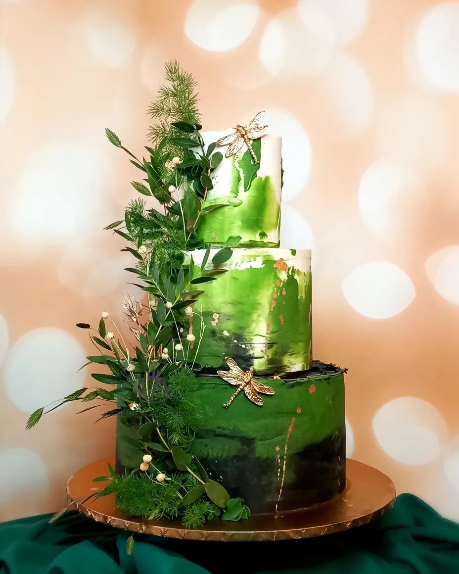 Trzypiętrowy tort w odcieniach zieleni, zdobiony żywymi roślinami i złotymi ważkami, ustawiony na złotej podstawie z zielonym materiałem w tle.