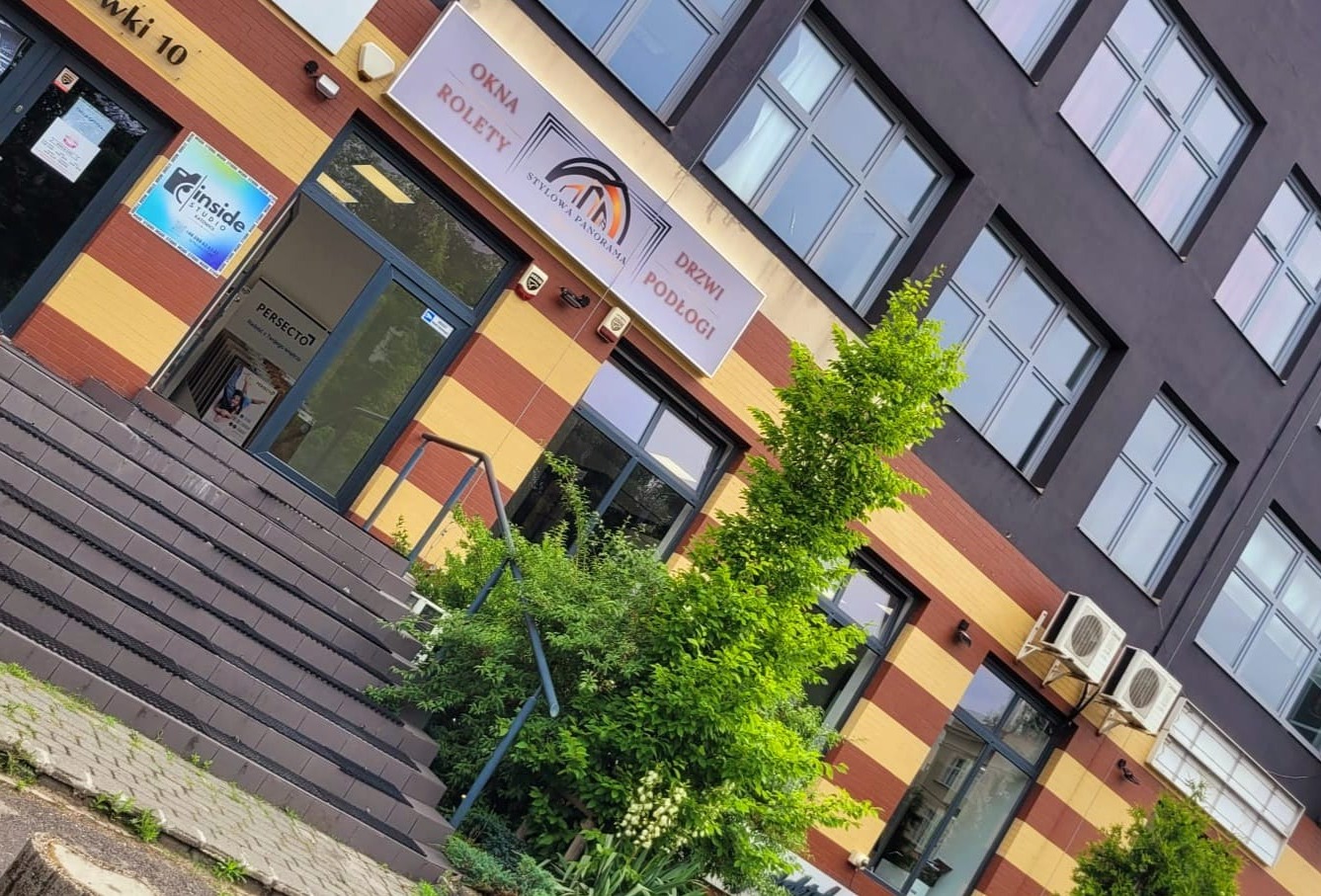 Fasada budynku z oknami, drzwiami i roletami, widoczny szyld firmy 'Stylowa Panorama' oferującej okna, rolety, drzwi i podłogi, zieleń przed wejściem.