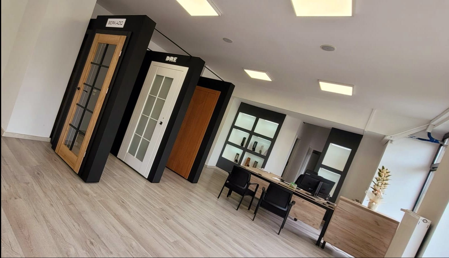 Wnętrze salonu z ekspozycją drzwi wewnętrznych różnych producentów (Berkado, DRE), ustawionych na podłodze wzdłuż ściany, widok na biurko obsługi klienta.