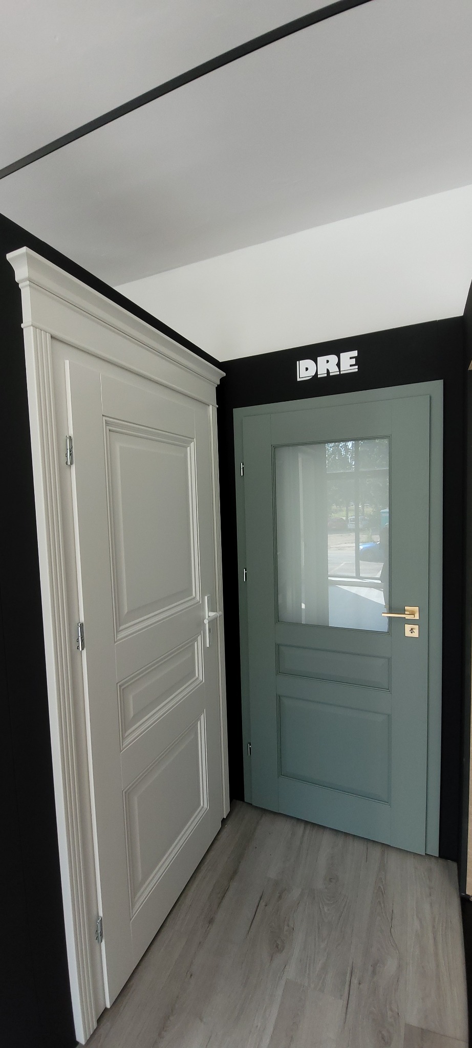 Dwie wewnętrzne drzwi: białe drzwi panelowe z ozdobną ramą i srebrną klamką oraz szaro-zielone drzwi z szybą i złotą klamką, na szarej podłodze, z logo DRE na czarnym tle.